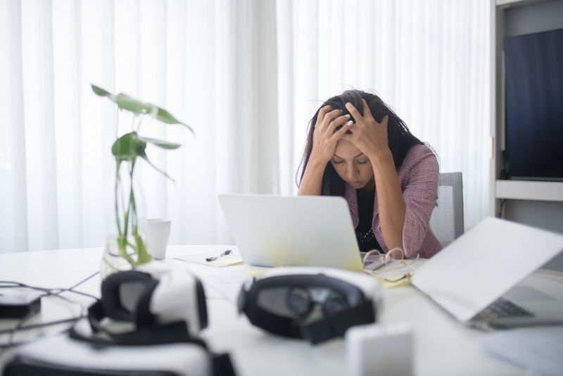 Mulher com as mãos na cabeça sentada em frente ao computador em ambiente de trabalho