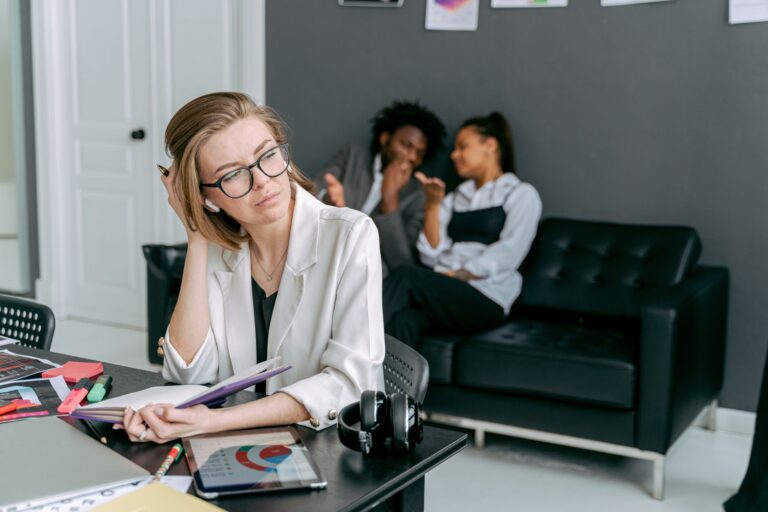 Mulher parece estressada em sua mesa de trabalho, com colegas ao fundo, ilustrando situação de assédio no ambiente de trabalho