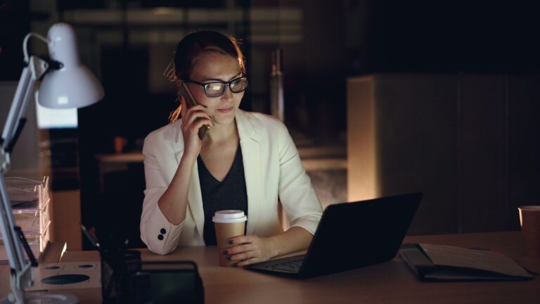 Mulher trabalhando à noite no notebook, com café e celular, em escritório