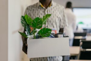 Homem carregando caixa com itens pessoais e uma planta ao deixar o escritório