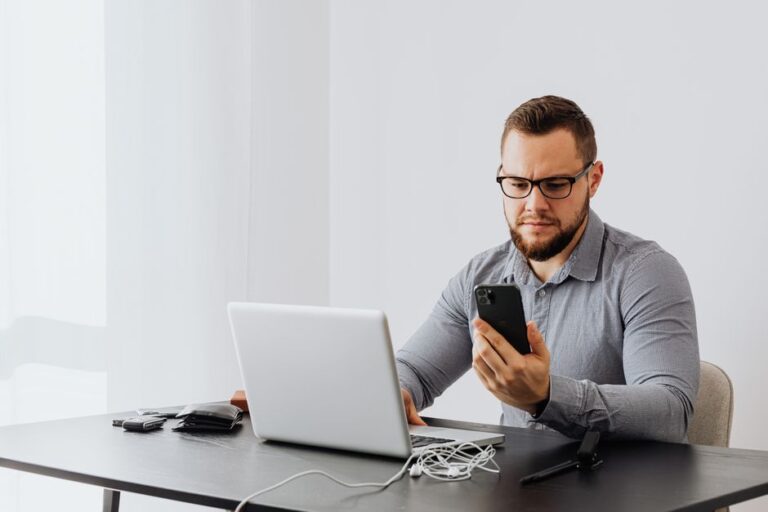 Trabalhador conferindo informações no celular e no notebook em uma mesa de escritório