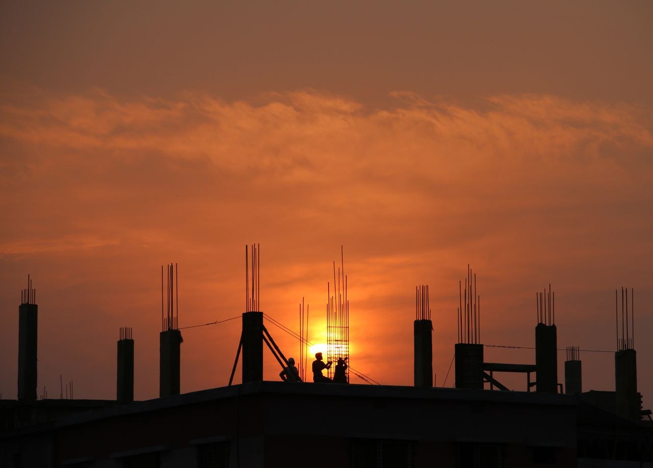 Silhueta de trabalhadores em obra ao entardecer, representando jornada longa