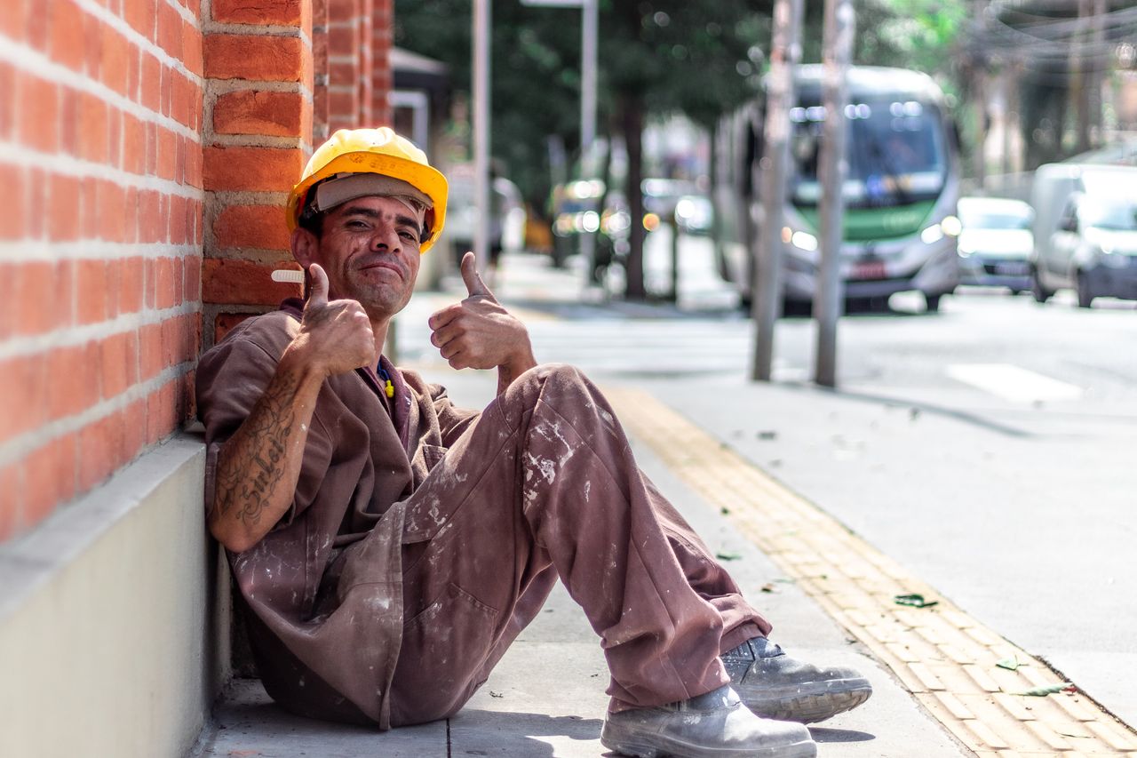 Trabalhador de capacete amarelo sentado na calçada fazendo sinal de positivo